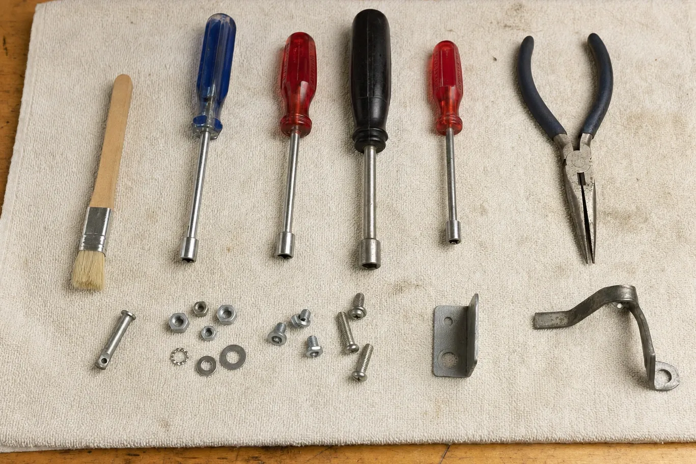 A bristle brush, four nut drivers and long-nose pliers laid out on a cloth over a wooden bench, with nuts, washers, screws and two small steel brackets set out below them