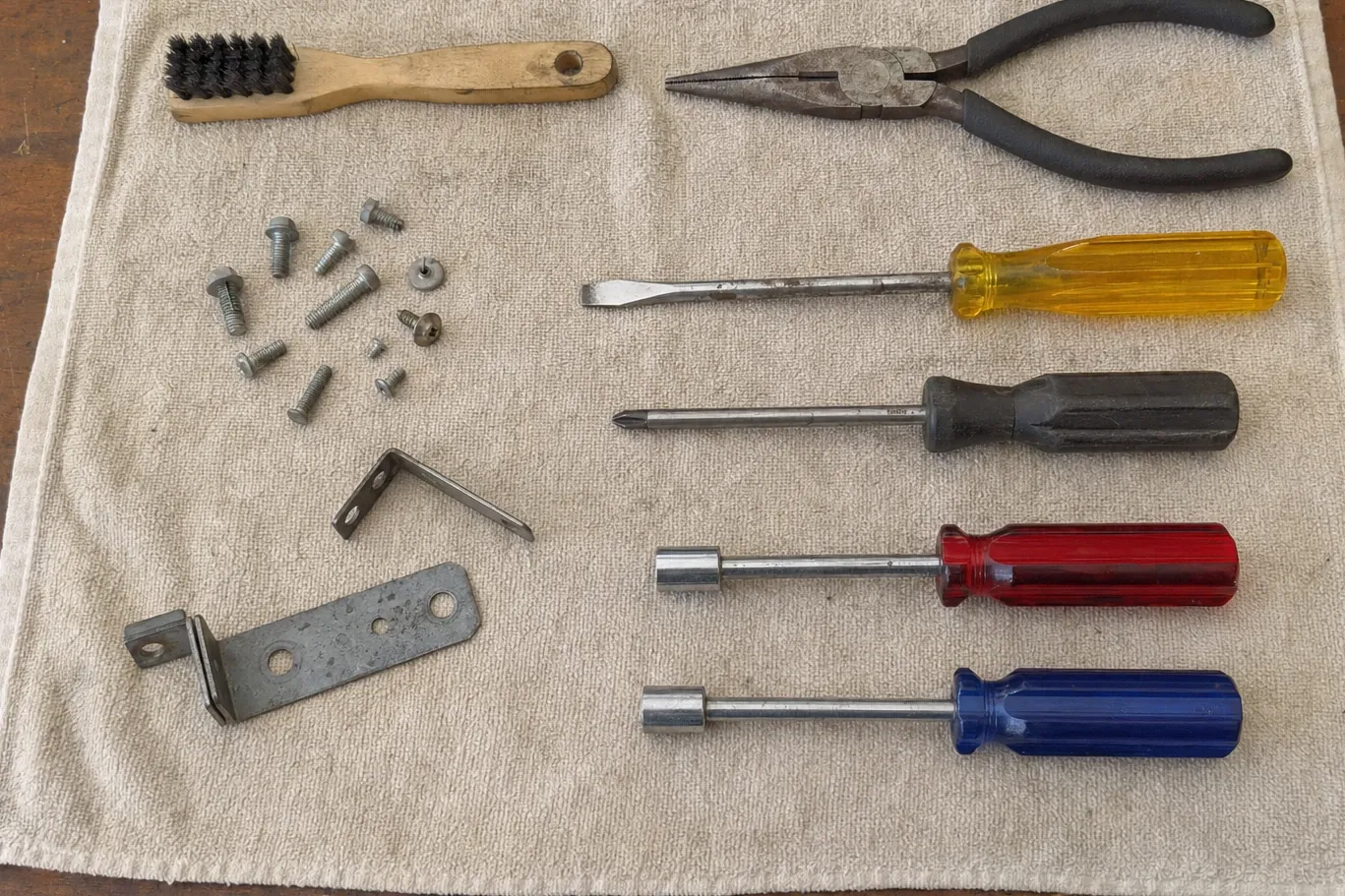 A wire brush, long-nose pliers, two screwdrivers and two nut drivers ranked on a cloth opposite a spread of machine screws, an angle bracket and a drilled flat bar